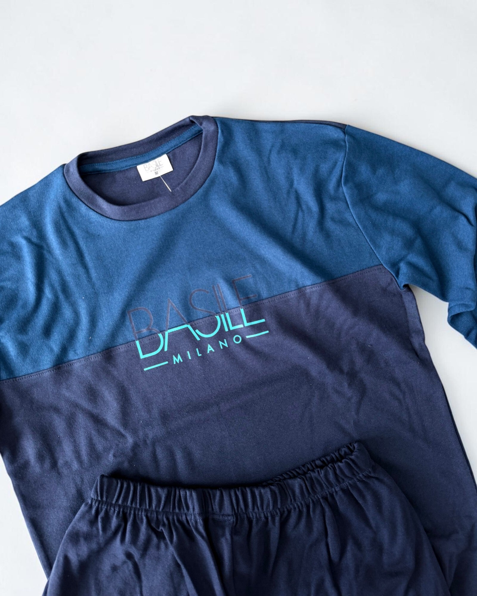 Dettaglio del girocollo e della stampa Basile Milano sulla maglia ottanio, con pantaloni blu scuro a vita elasticizzata