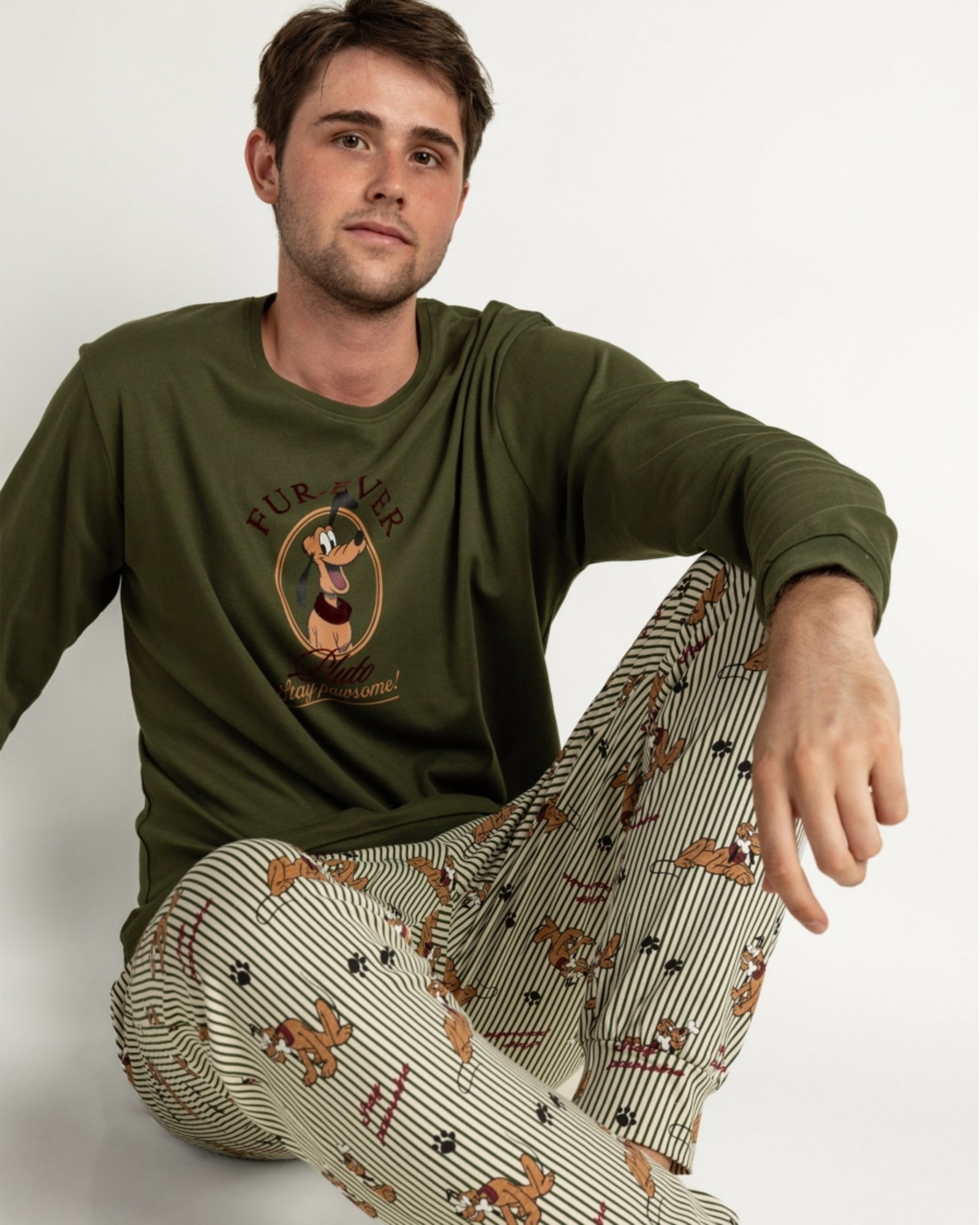 Modello seduto con pigiama Disney Pluto 64928: maglia verde con stampa frontale e pantaloni a righe con Pluto e impronte.