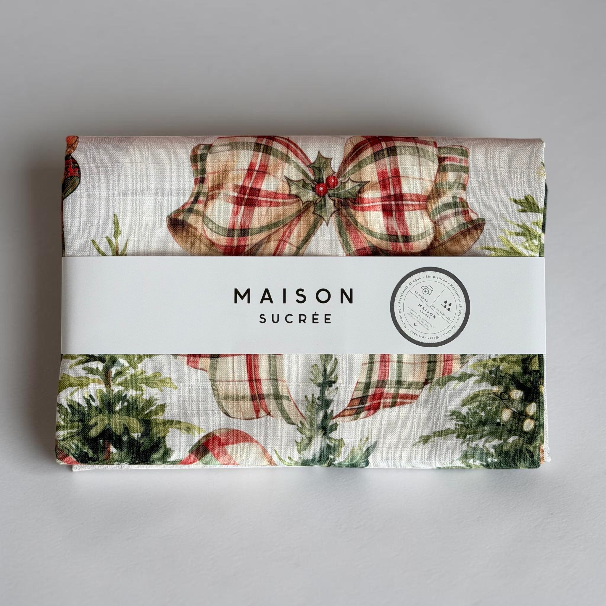 Tovaglia Natale in salotto ripiegata con fascetta Maison Sucrée e dettaglio di un fiocco tartan sopra la fantasia di abeti.